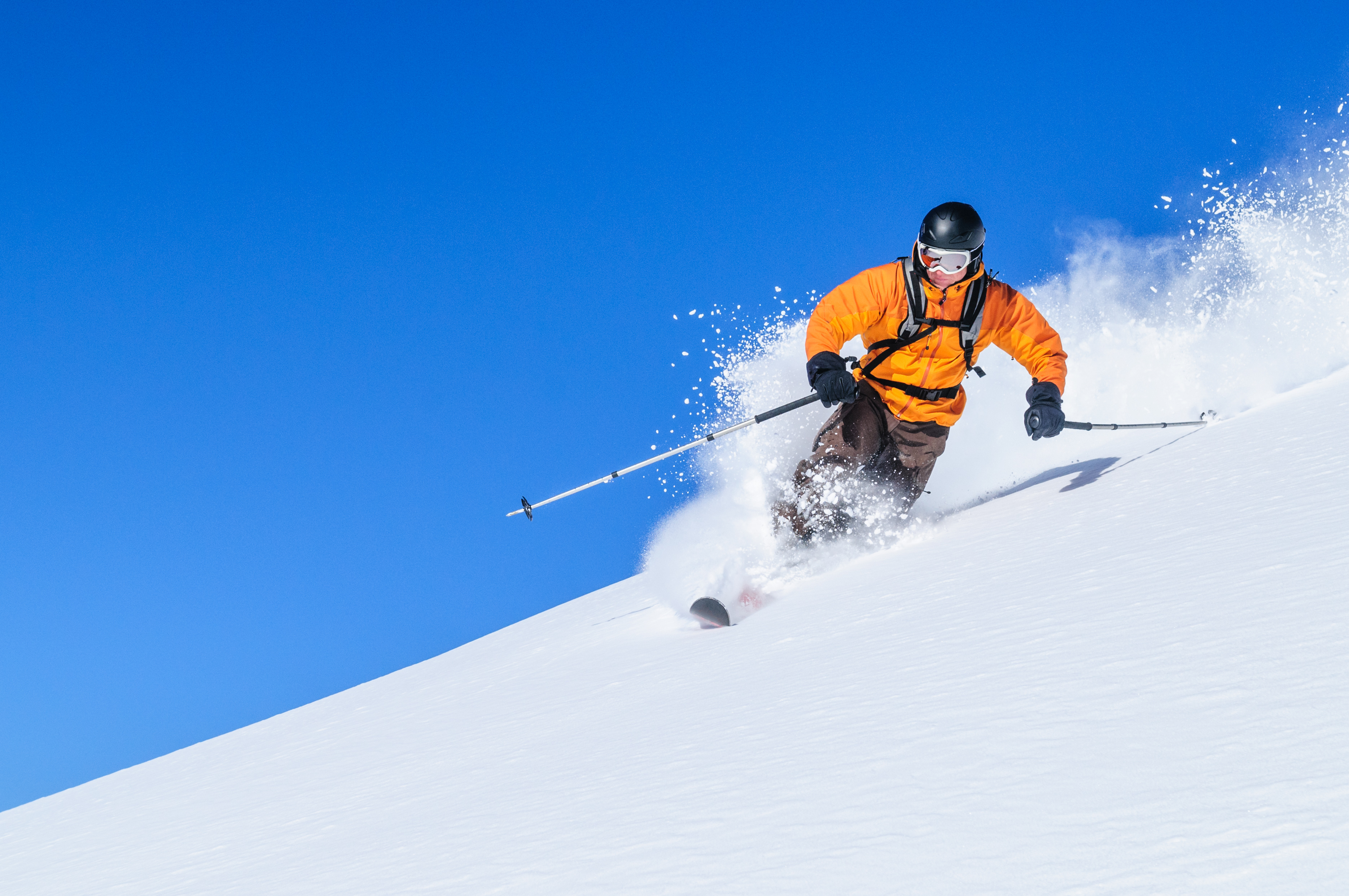 Skieur en veste orange dévalant une piste enneigée, soulevant de la poudreuse sous un ciel bleu vif.