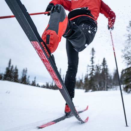 Skieur de fond en pleine action avec un pied levé, portant des bottes rouges et des skis Atomic noirs, projetant de la neige sur une piste.
