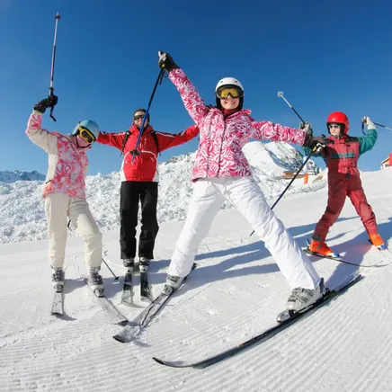 Quatre skieurs, bras levés et souriants, posent sur une piste ensoleillée avec des montagnes enneigées et un ciel bleu clair.