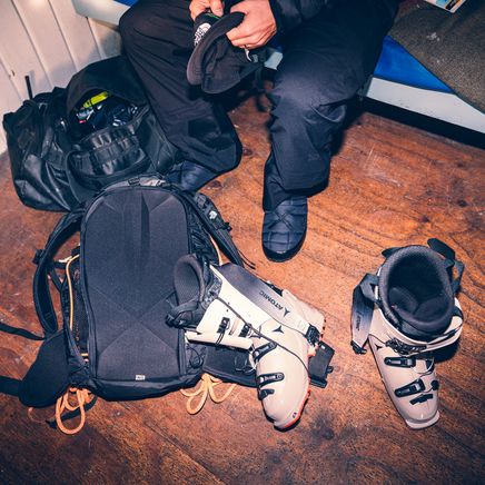 Un skieur se prépare avec son équipement : un casque Monster Energy, des bottes de ski, des sacs à dos sur un plancher en bois.