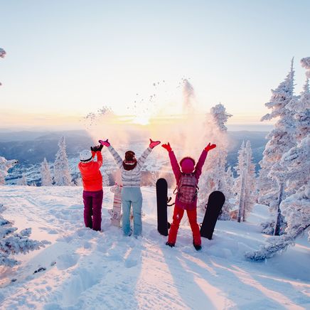 Trois personnes sur une montagne enneigée, jetant de la poudreuse en l'air sous un soleil doré. Arbres givrés et snowboards.
