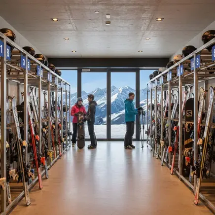 Salle de rangement de skis et casques. Trois personnes se préparent, avec une vue imprenable sur les montagnes enneigées par une grande baie vitrée.