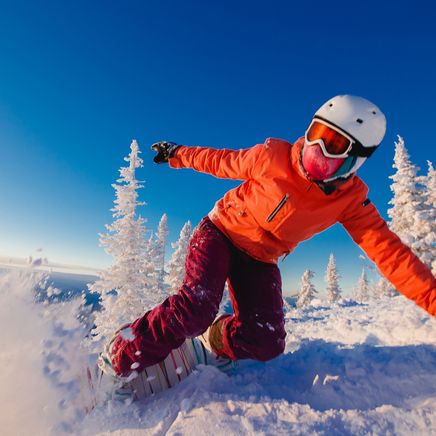 Snowboarder en veste orange dévalant une montagne enneigée, projetant de la poudreuse sous un ciel bleu avec des arbres givrés.