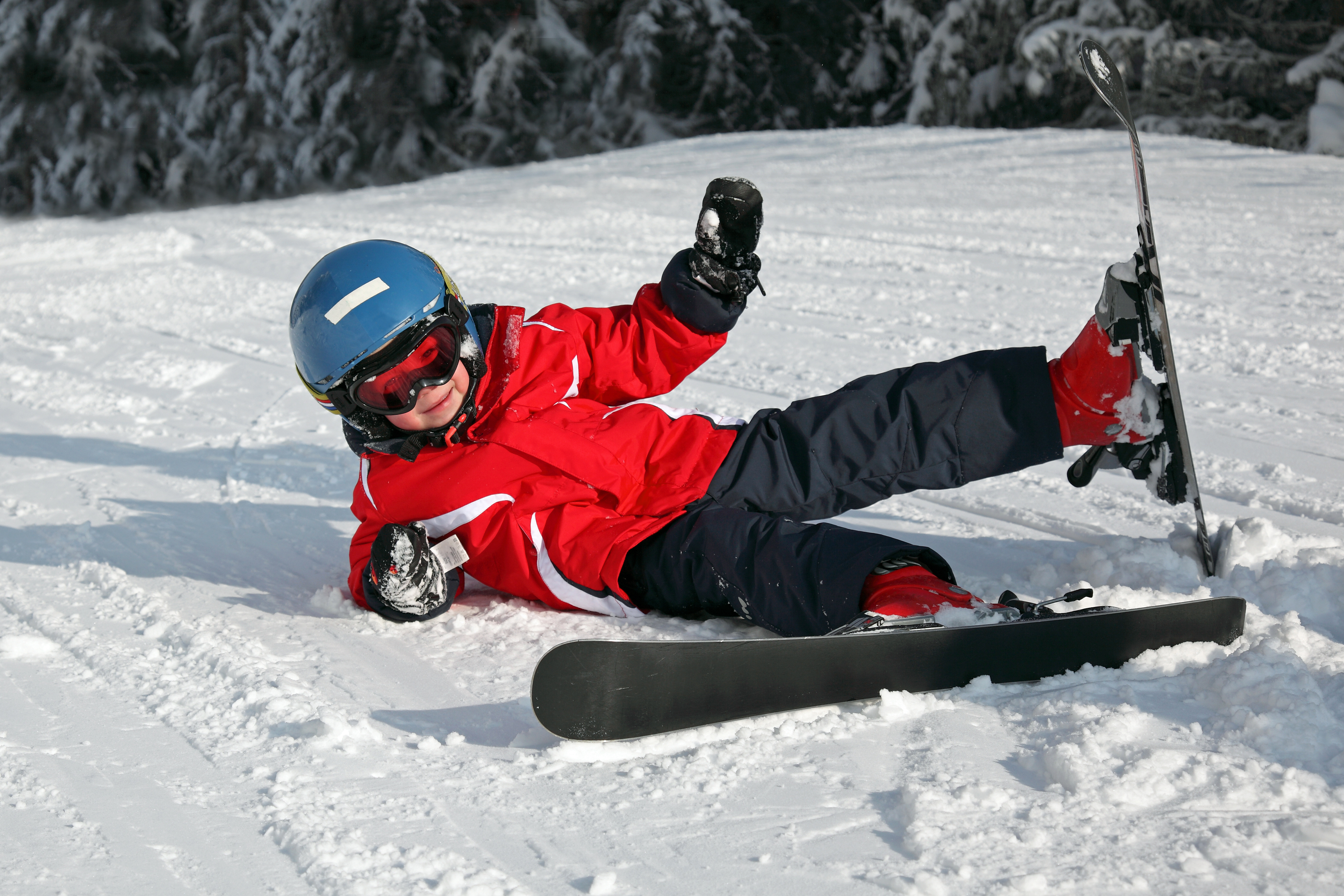 Un enfant en veste rouge et casque bleu est tombé à ski et sourit, allongé dans la neige sur une piste enneigée.
