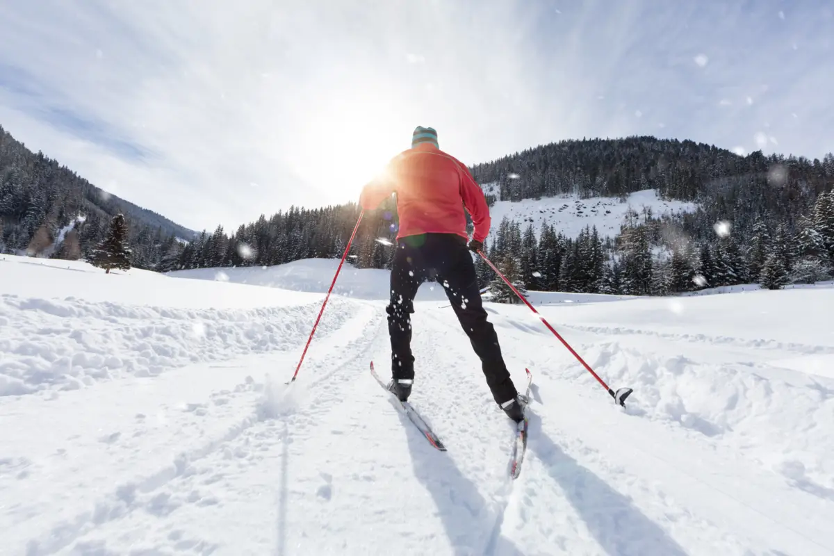 Skieur en vêtements rouges pratiquant le ski de fond sur une piste enneigée, entourée de montagnes et de forêts sous un ciel lumineux.