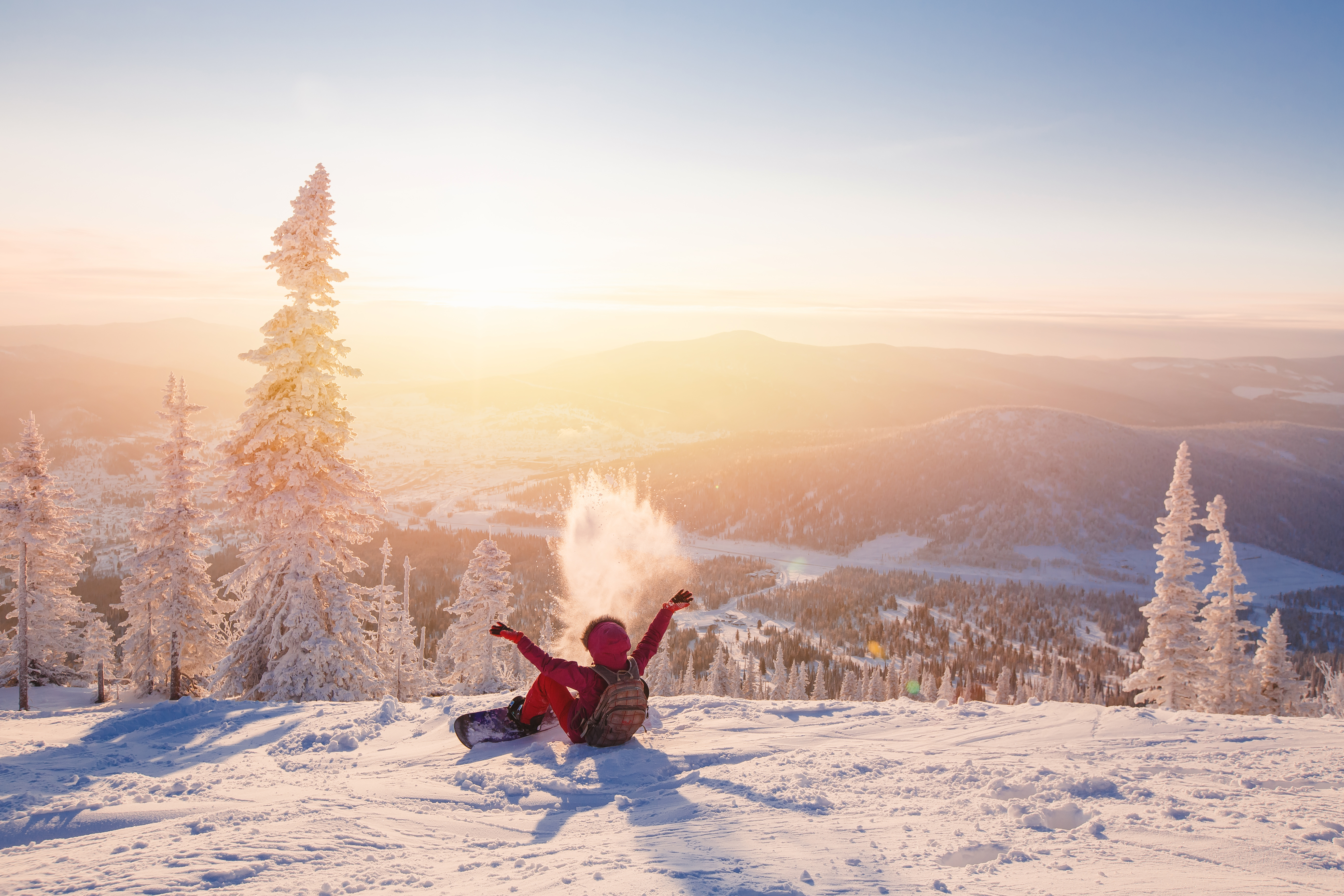 Snowboarder assis sur un sommet enneigé au lever du soleil, lançant de la neige. Sapins givrés et montagnes à l'horizon.