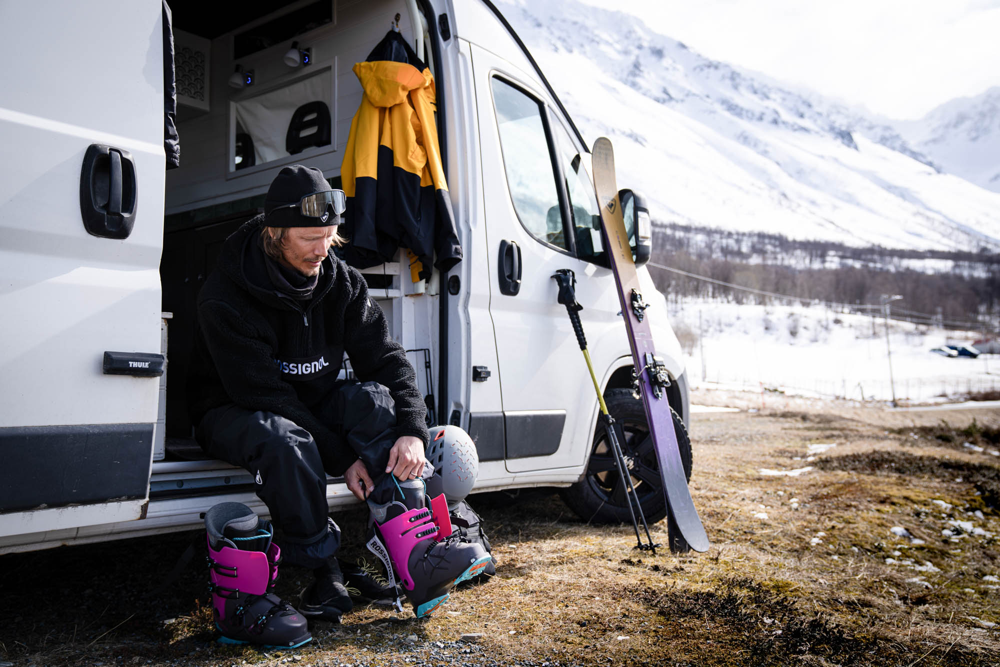 Un homme barbu met ses chaussures de ski roses et noires sur le pas de son van, des skis appuyés, face à des montagnes enneigées.