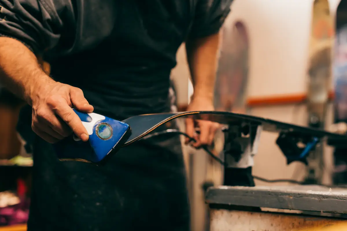 Gros plan sur des mains préparant une planche de snowboard ou de ski avec un outil bleu sur sa base sombre, dans un atelier.
