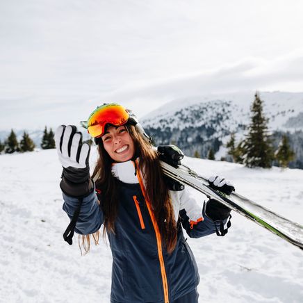 Femme souriante en tenue de ski, skis sur l'épaule, saluant sur une montagne enneigée.