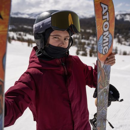 Jeune skieur avec casque et lunettes, tenant des skis Atomic colorés sur une piste enneigée avec montagnes en arrière-plan.