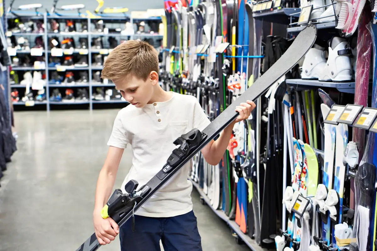 Jeune garçon dans un magasin de sport, tenant et examinant un ski. Des rayons remplis d'autres skis et chaussures de ski sont en arrière-plan.