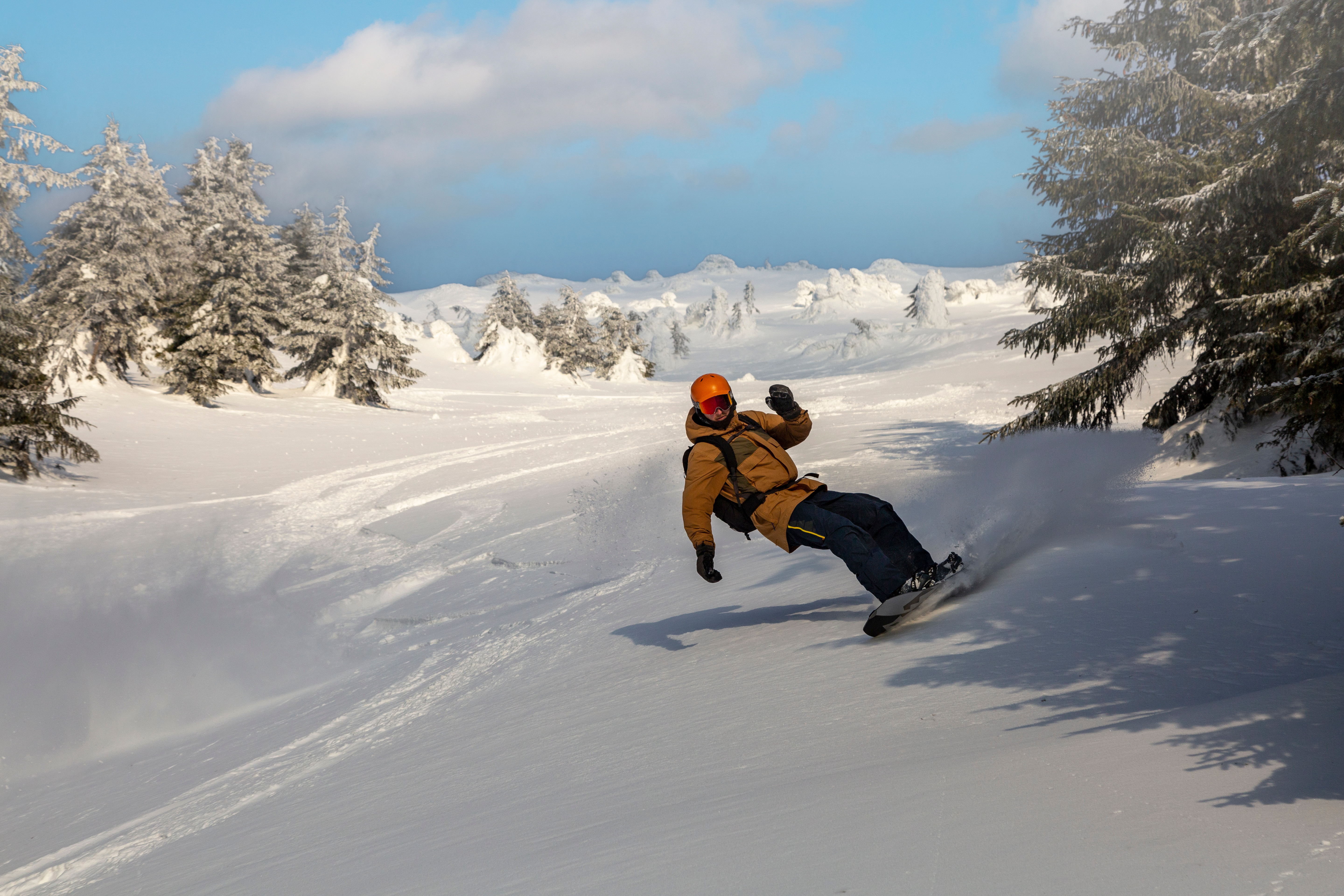 Un snowboarder en veste marron et casque orange tourne dans la neige fraîche sur une pente enneigée. Le soleil brille, des sapins givrés sont visibles.