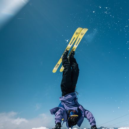 Skieur en poirier sur la neige avec des skis jaunes Rossignol pointant vers le ciel bleu éclatant, des flocons scintillent.