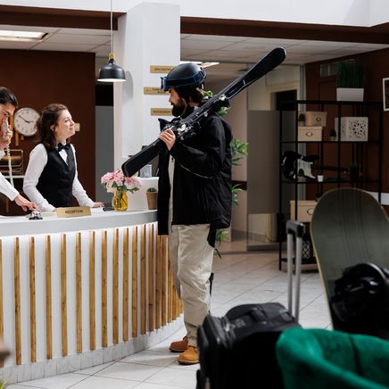 Homme avec des skis à la réception d'un hôtel, assisté par deux employés. Une skieuse attend, regardant son téléphone.