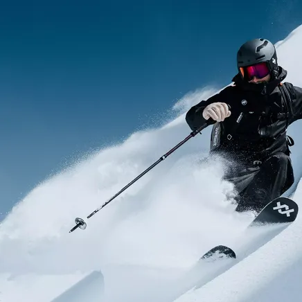 Skieur en noir descendant une pente de poudreuse sous un ciel bleu, soulevant un nuage de neige.