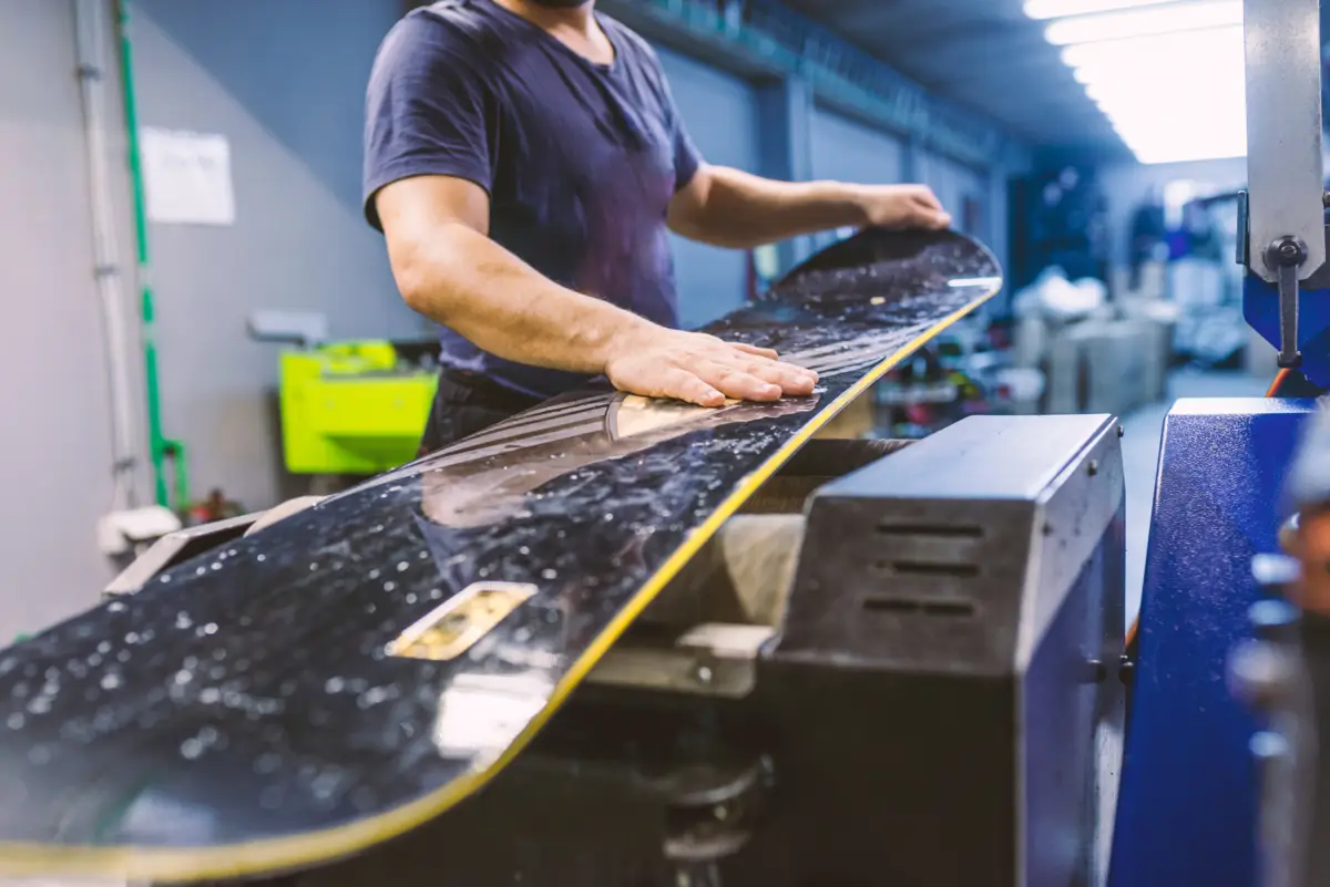 Un homme prépare la semelle d'un snowboard noir et jaune sur une machine de maintenance en atelier.