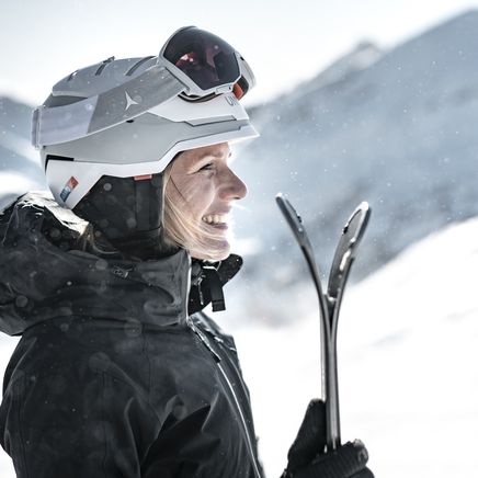 Femme blonde souriante en casque de ski blanc et veste noire, tenant des skis, dans un paysage de montagne enneigé et ensoleillé.