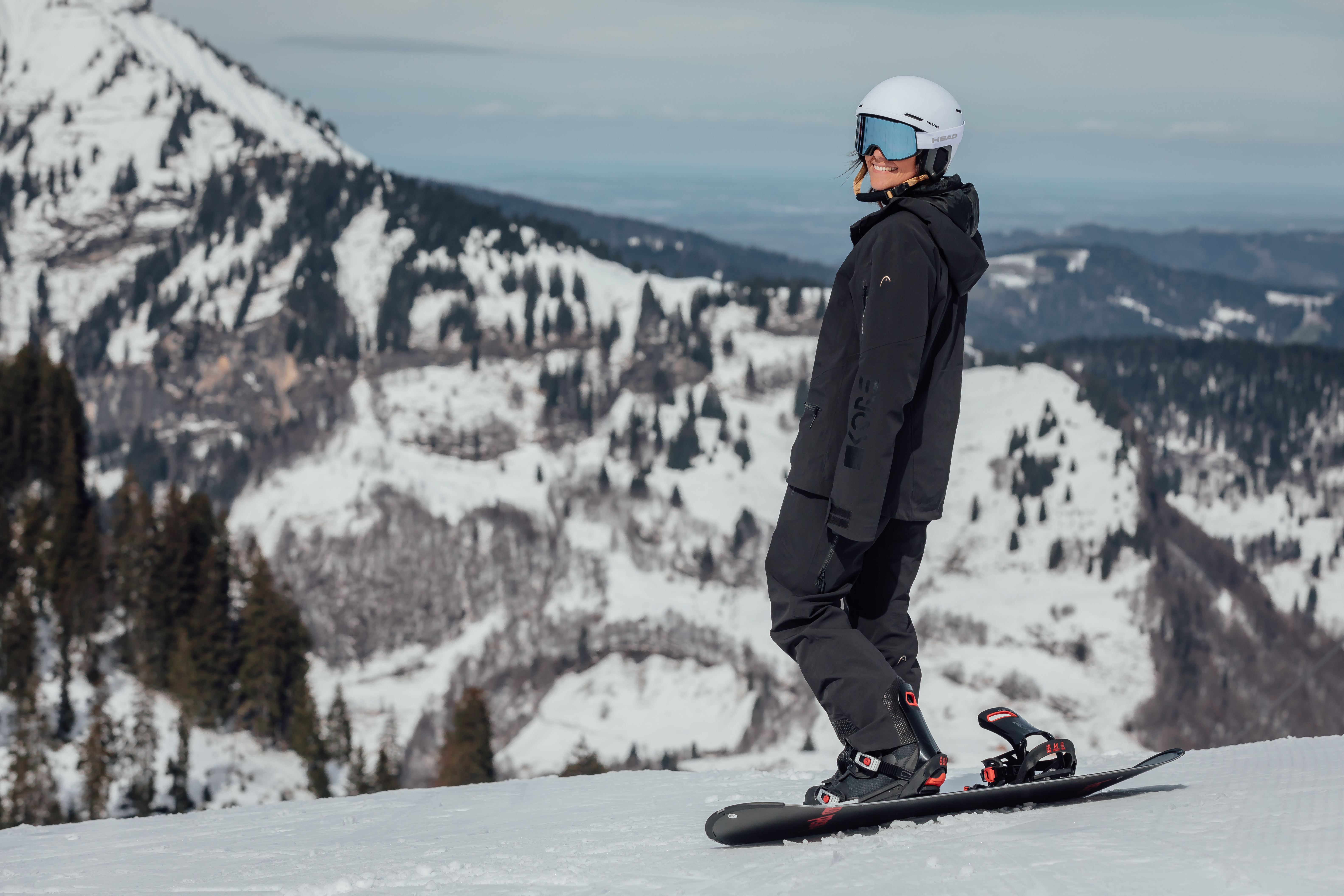 Une personne en tenue de snowboard noire avec casque blanc et lunettes bleues, debout sur sa planche sur une piste de montagne enneigée.