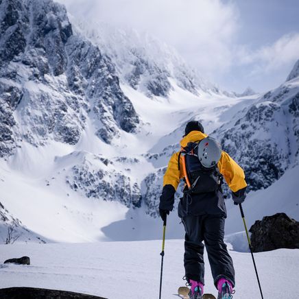 Skieur en veste jaune faisant du ski de randonnée dans une vallée enneigée, entouré de montagnes majestueuses sous un ciel nuageux.