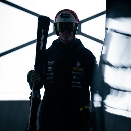 Skieur professionnel en tenue sombre, casque rouge et lunettes, tenant ses skis, silhouetté devant une fenêtre lumineuse.
