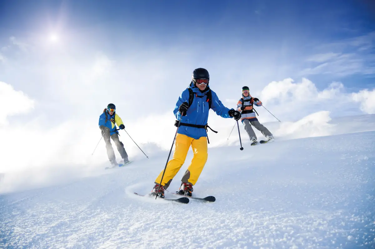 Trois skieurs dévalent une pente enneigée sous un ciel bleu ensoleillé. Le skieur principal est en veste bleue et pantalon jaune.