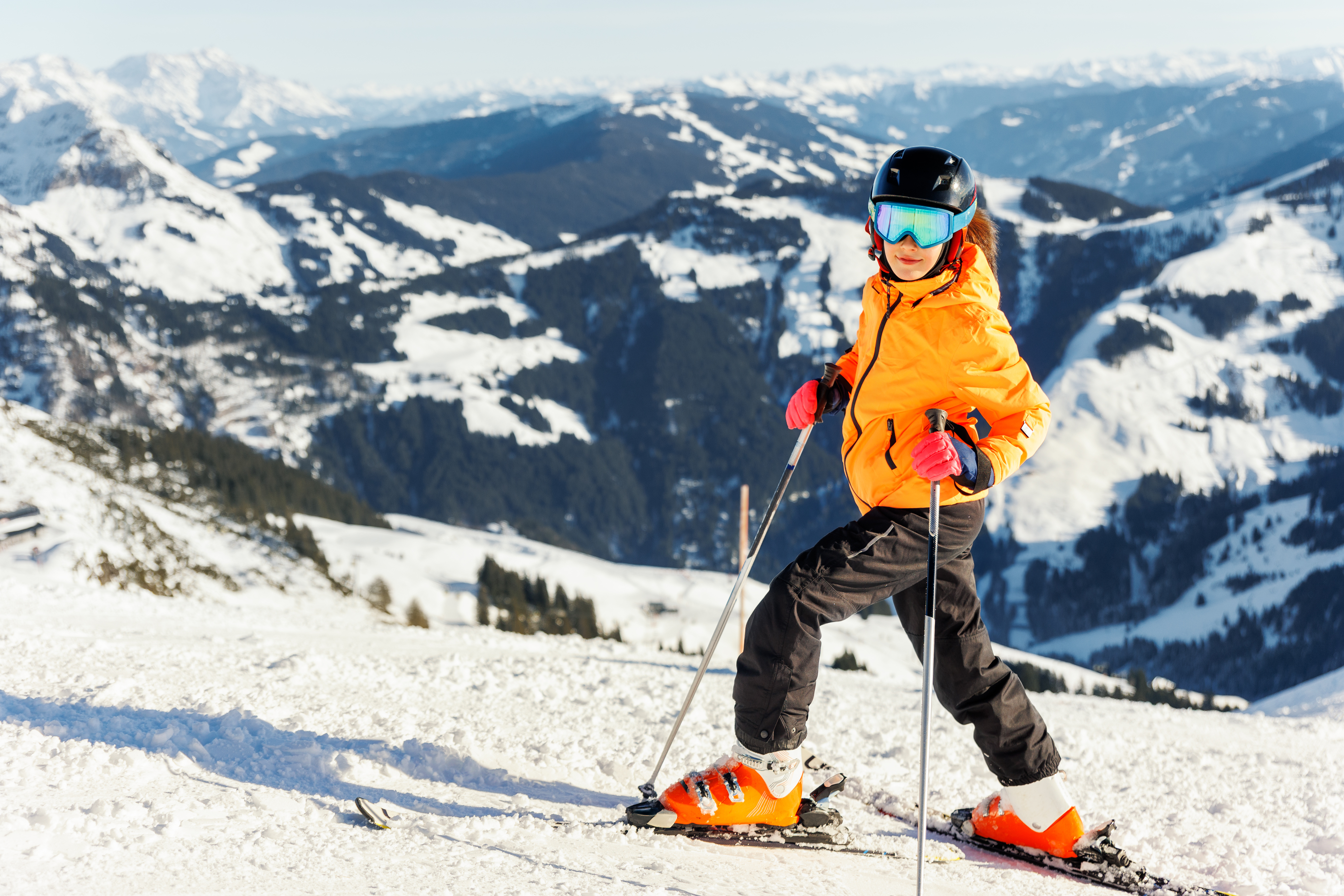 Enfant skieur en veste orange, casque et lunettes bleues sur une piste enneigée avec des montagnes en arrière-plan sous un ciel ensoleillé.
