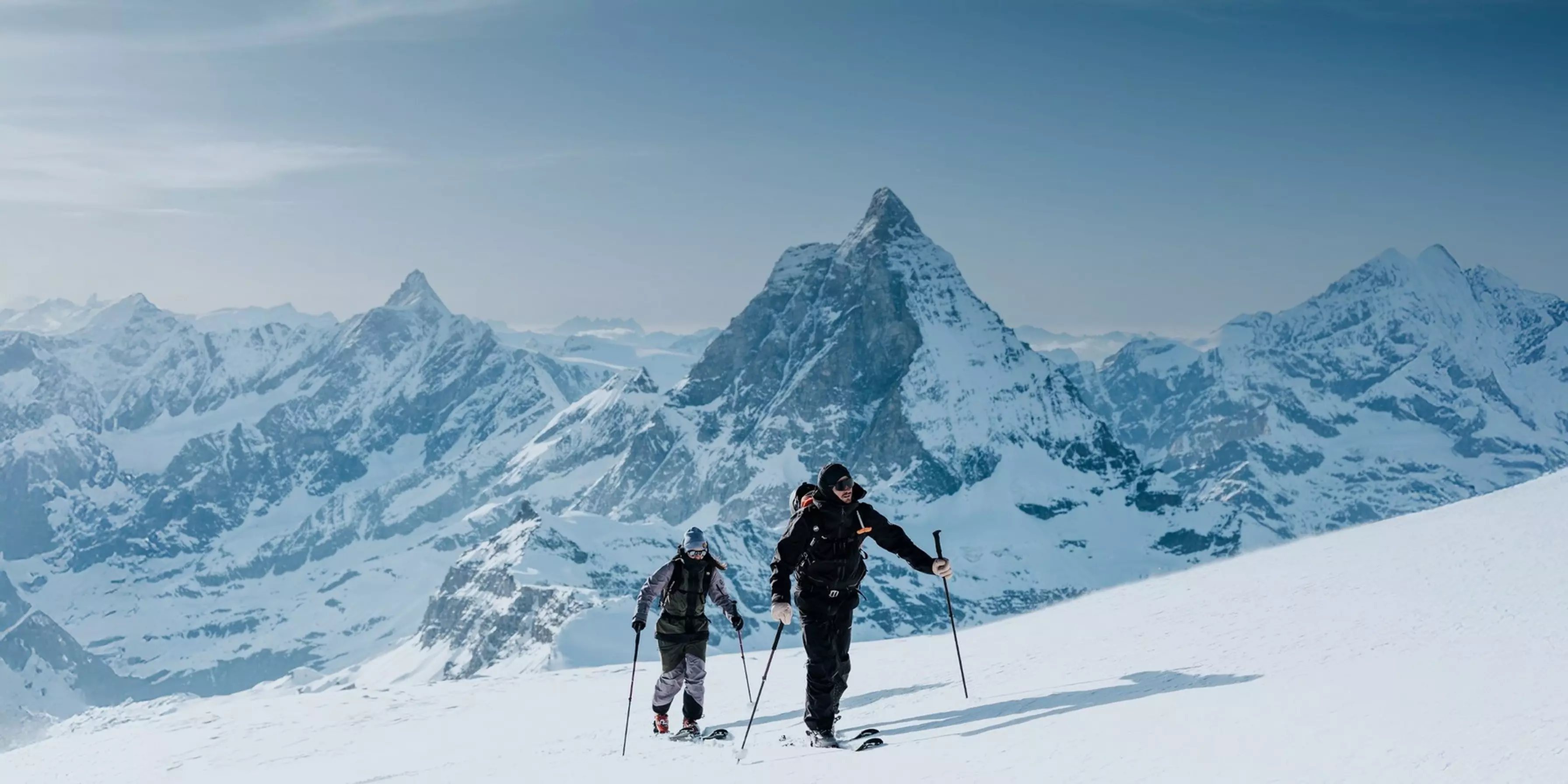 Deux randonneurs, l'un à ski, l'autre en raquettes, montent une pente enneigée face à de vastes montagnes sous un ciel clair.