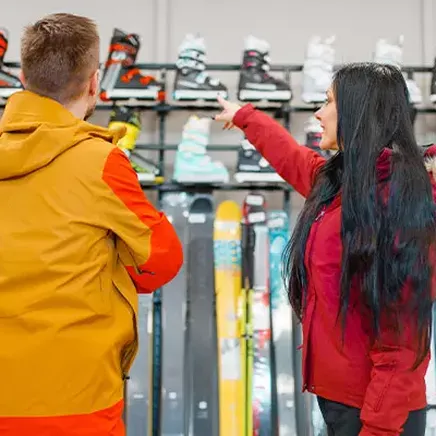 Dans un magasin de ski, une femme en doudoune rouge pointe du doigt des chaussures à un homme, entourés de skis et bottes.