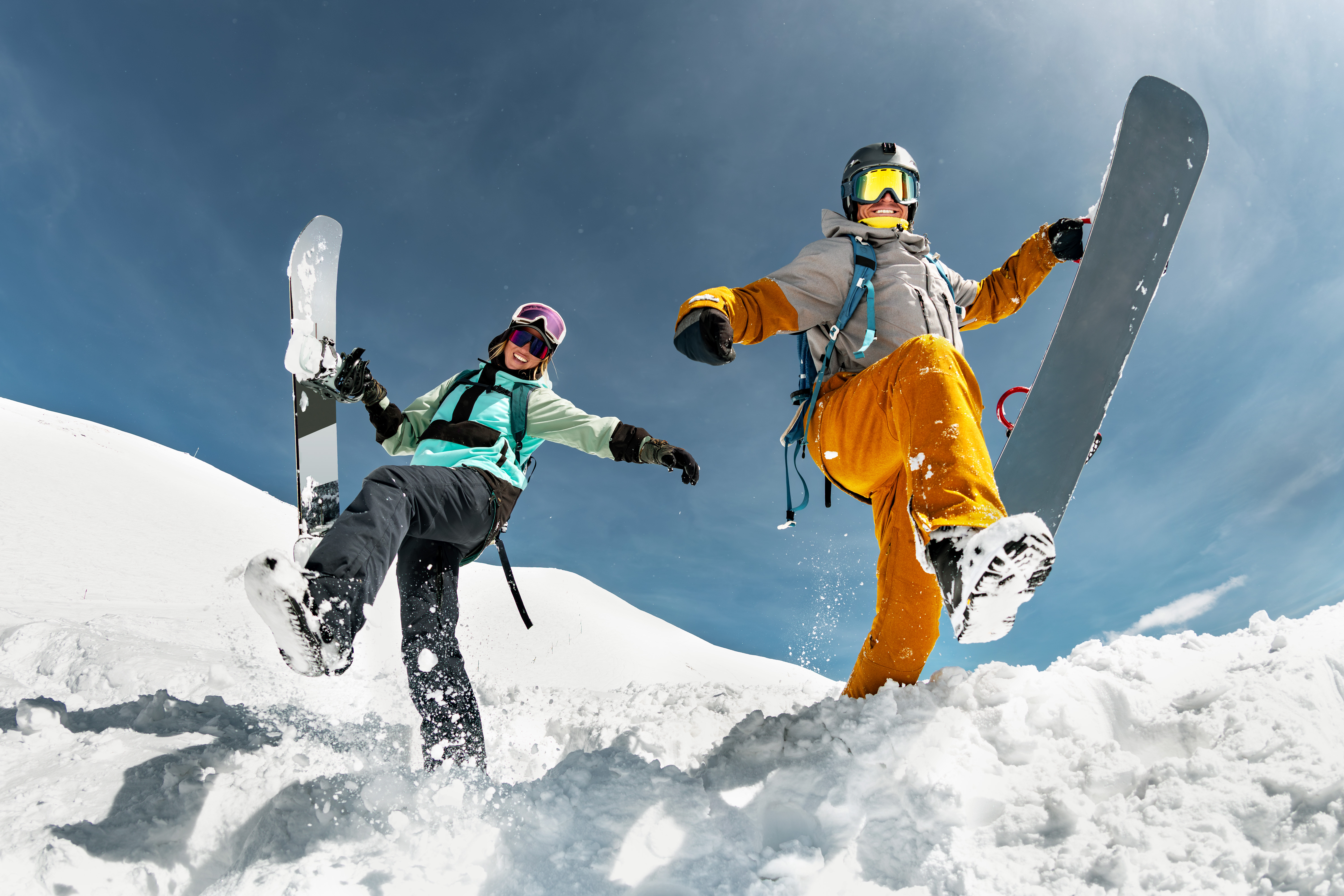 Deux snowboarders joyeux, un homme et une femme, posent avec leurs planches dans la neige poudreuse sous un ciel bleu.
