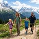 Famille souriante en randonnée sur un sentier de montagne, entourée de sommets enneigés, de forêts verdoyantes et de fleurs colorées sous un ciel bleu.