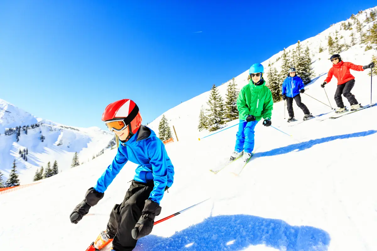 Des skieurs, dont un enfant en bleu au premier plan, descendent une piste enneigée sous un ciel clair et ensoleillé.