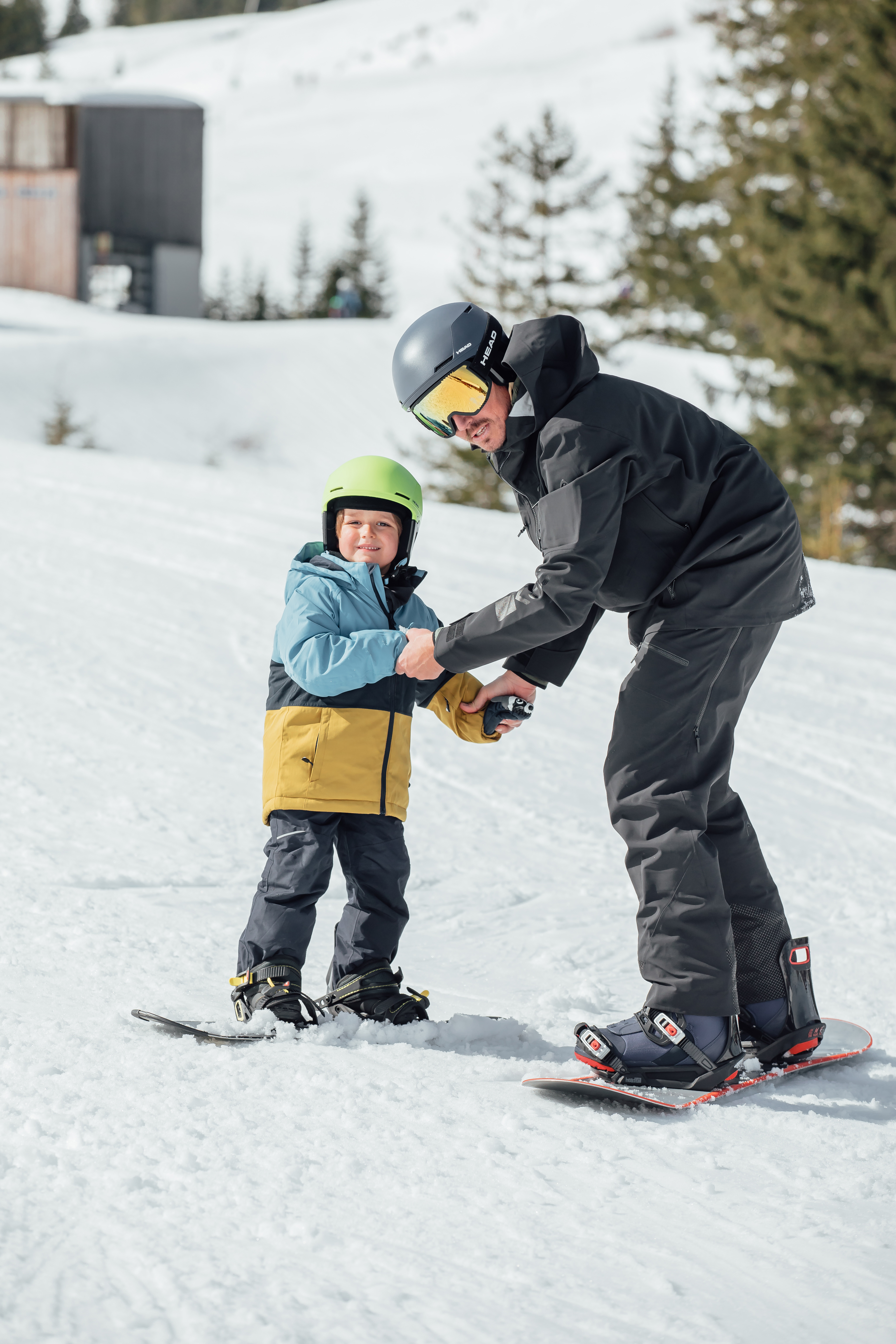 Un homme en snowboard aide un enfant souriant à faire de la planche sur une piste enneigée.