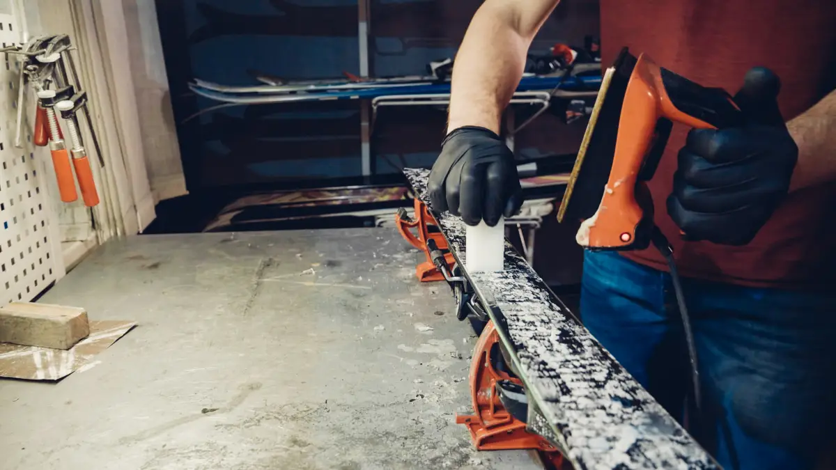 Personne en gants noirs appliquant de la cire sur un ski avec un fer à farter orange.