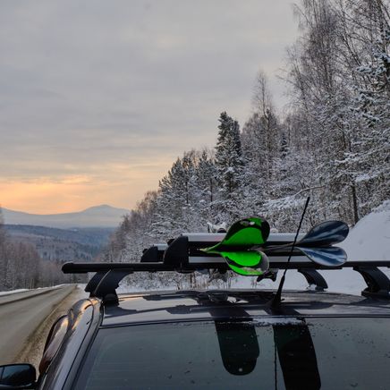 Voiture avec pagaies sur le toit, sur une route enneigée bordée d'une forêt hivernale, sous un ciel nuageux avec des montagnes lointaines.