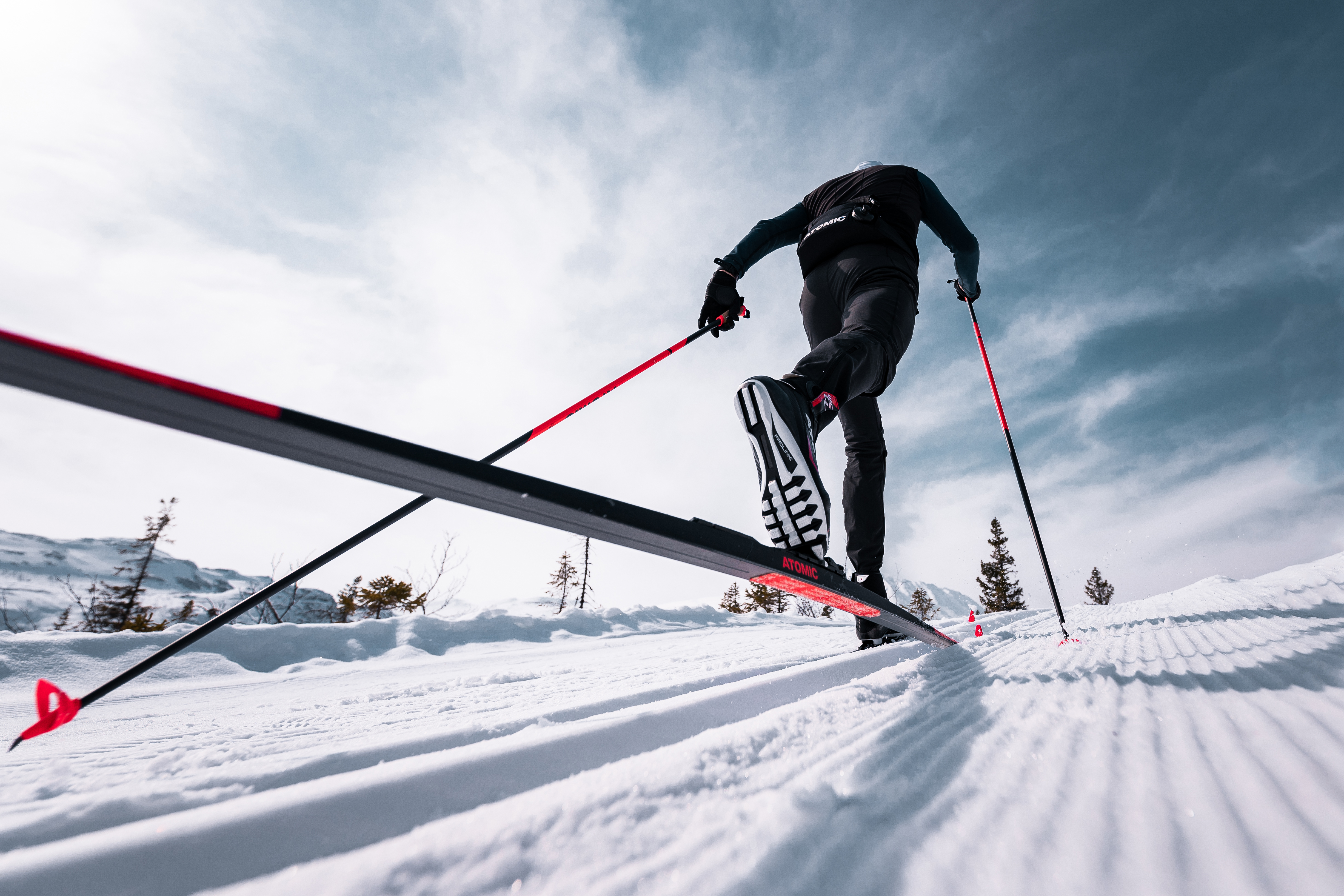 Vue en contre-plongée d'un skieur de fond montant une piste enneigée avec des skis et des bâtons de ski rouges, sous un ciel nuageux.