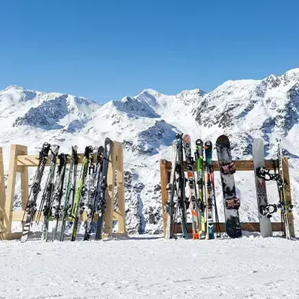 Nombreux skis et snowboards de toutes couleurs dans des râteliers en bois sur la neige, devant des montagnes enneigées sous un ciel bleu.