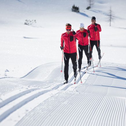 Trois hommes en vestes rouges font du ski de fond sur une piste enneigée et damée, sous un soleil éclatant.
