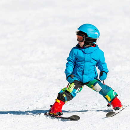 Jeune enfant en tenue de ski bleue et colorée, casque et lunettes, apprenant à skier sur une piste enneigée.