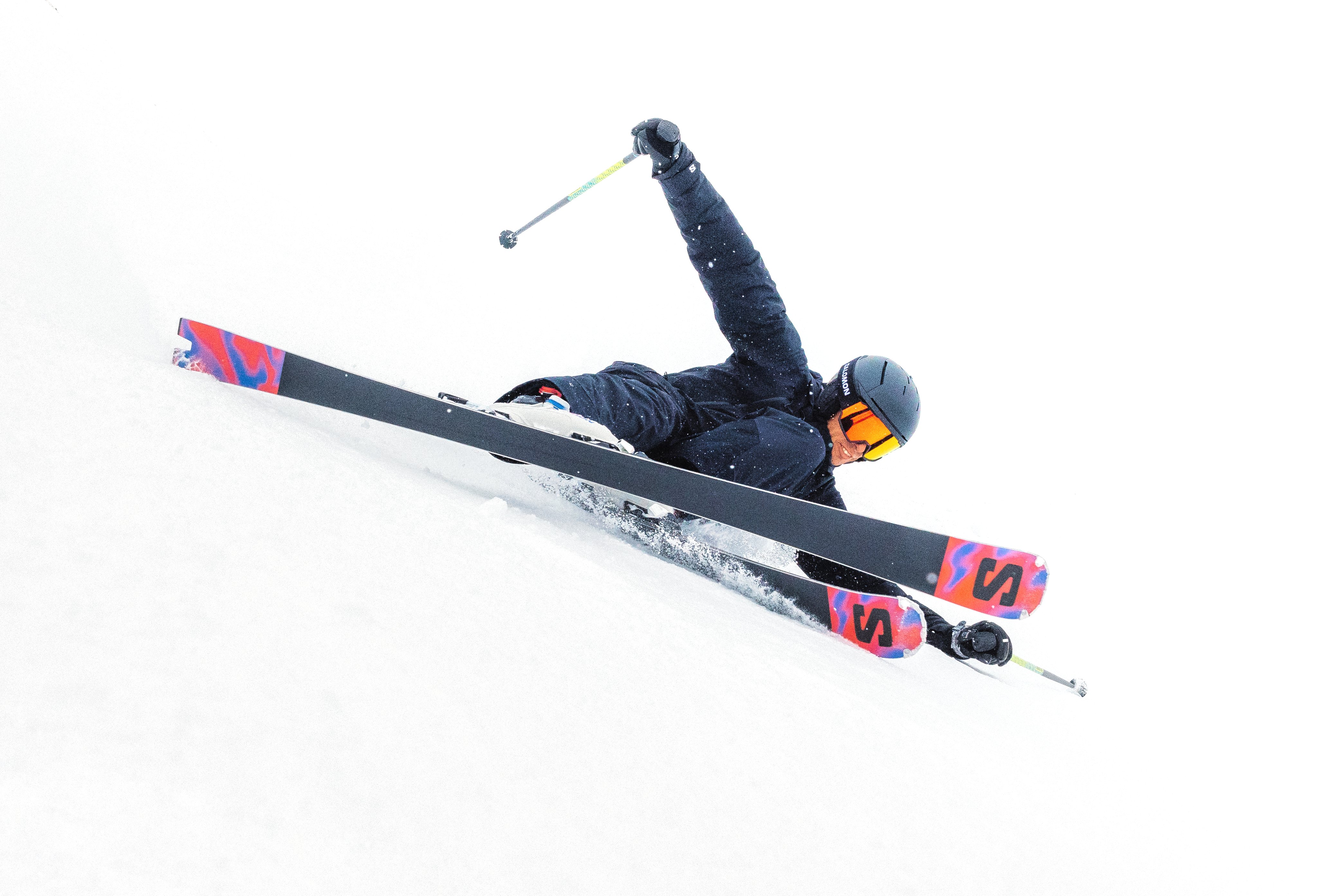 Un skieur en tenue noire, casque et lunettes, réalise un virage extrême sur une pente enneigée, skis Salomon roses et noirs, presque couché sur le flanc.