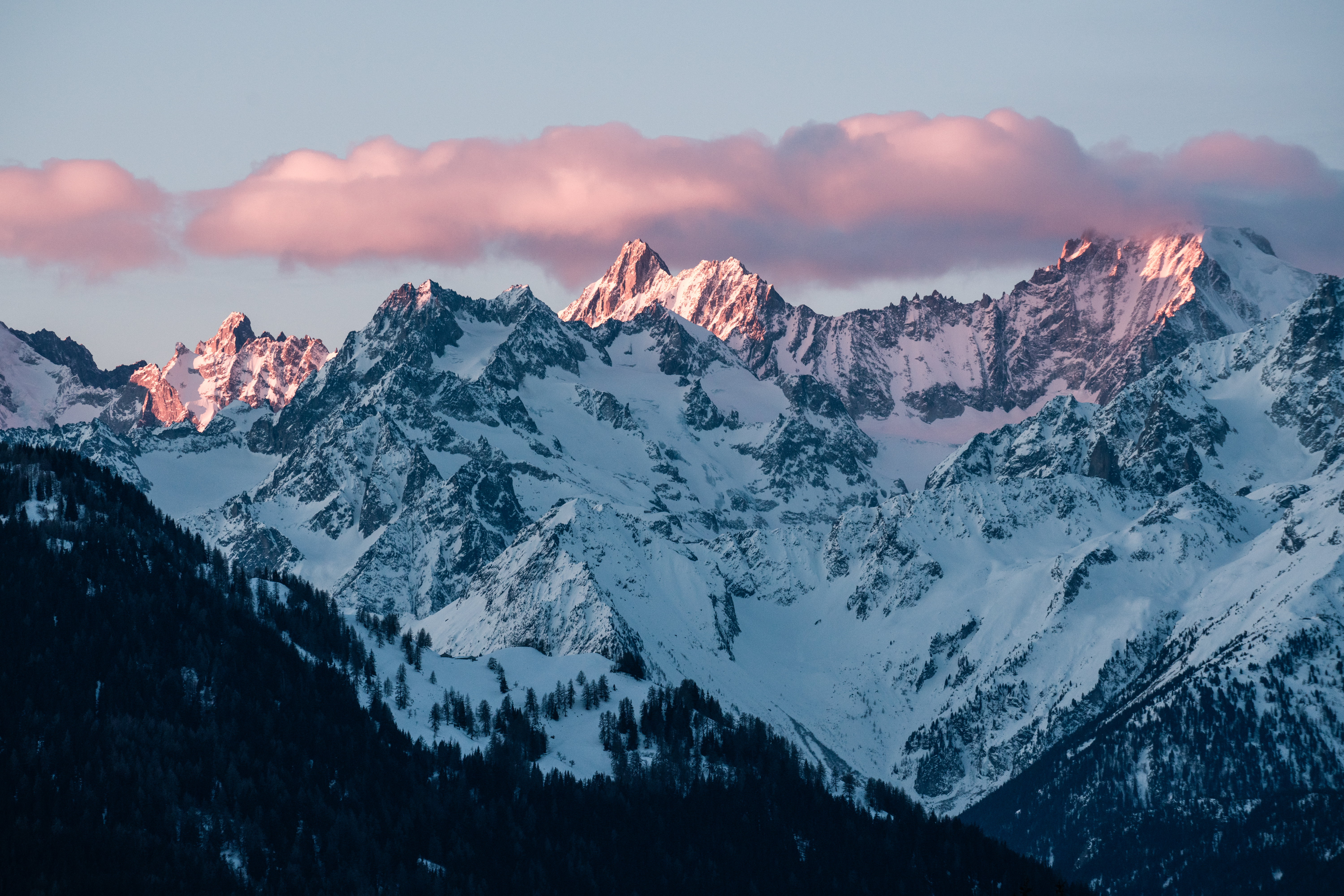 Montagnes enneigées illuminées par une douce lumière rosée sur les sommets et les nuages, avec des forêts sombres en contrebas.