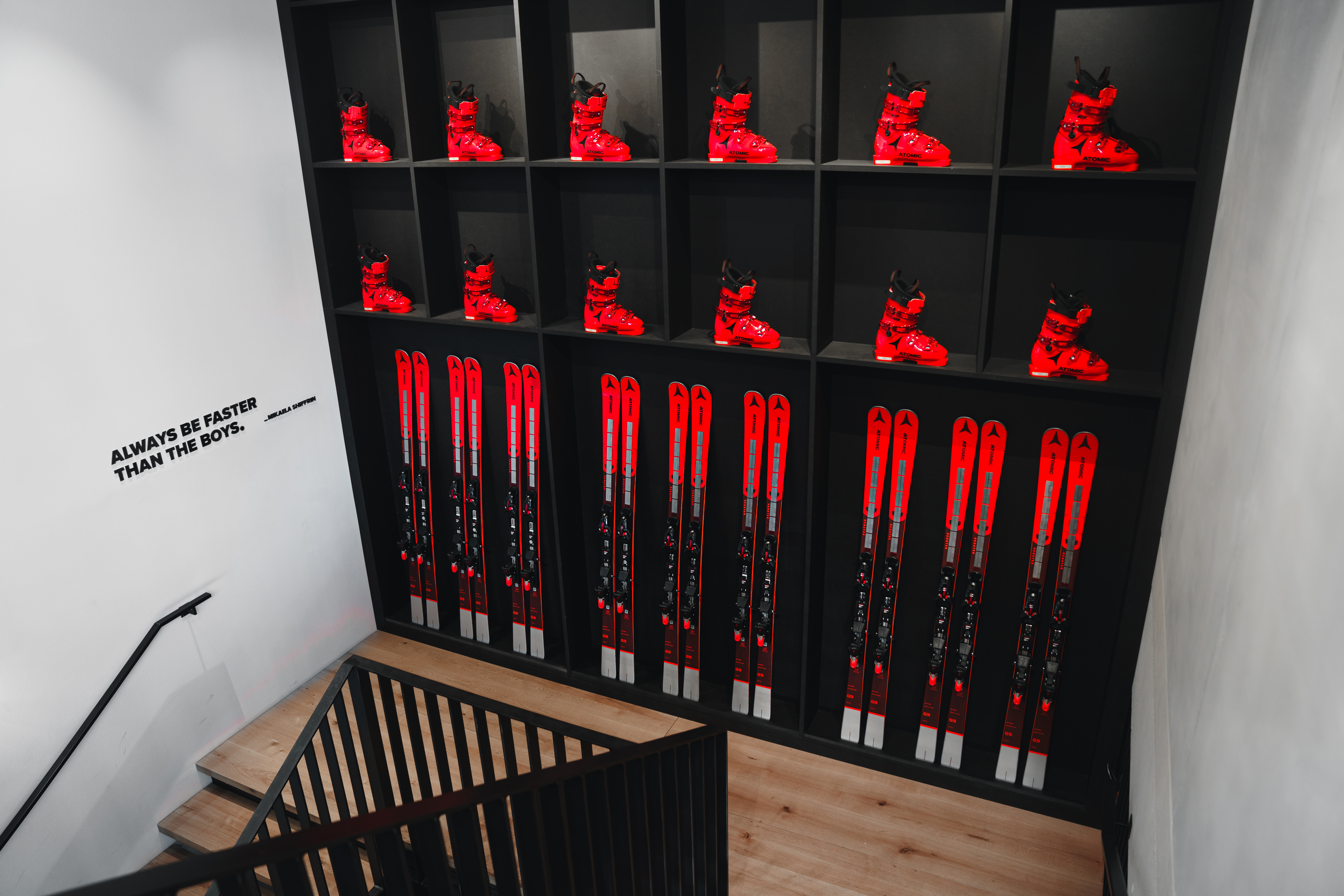 Bottes et skis de ski rouges alignés sur des étagères noires, avec la citation "ALWAYS BE FASTER THAN THE BOYS" sur un mur blanc à côté d'un escalier.